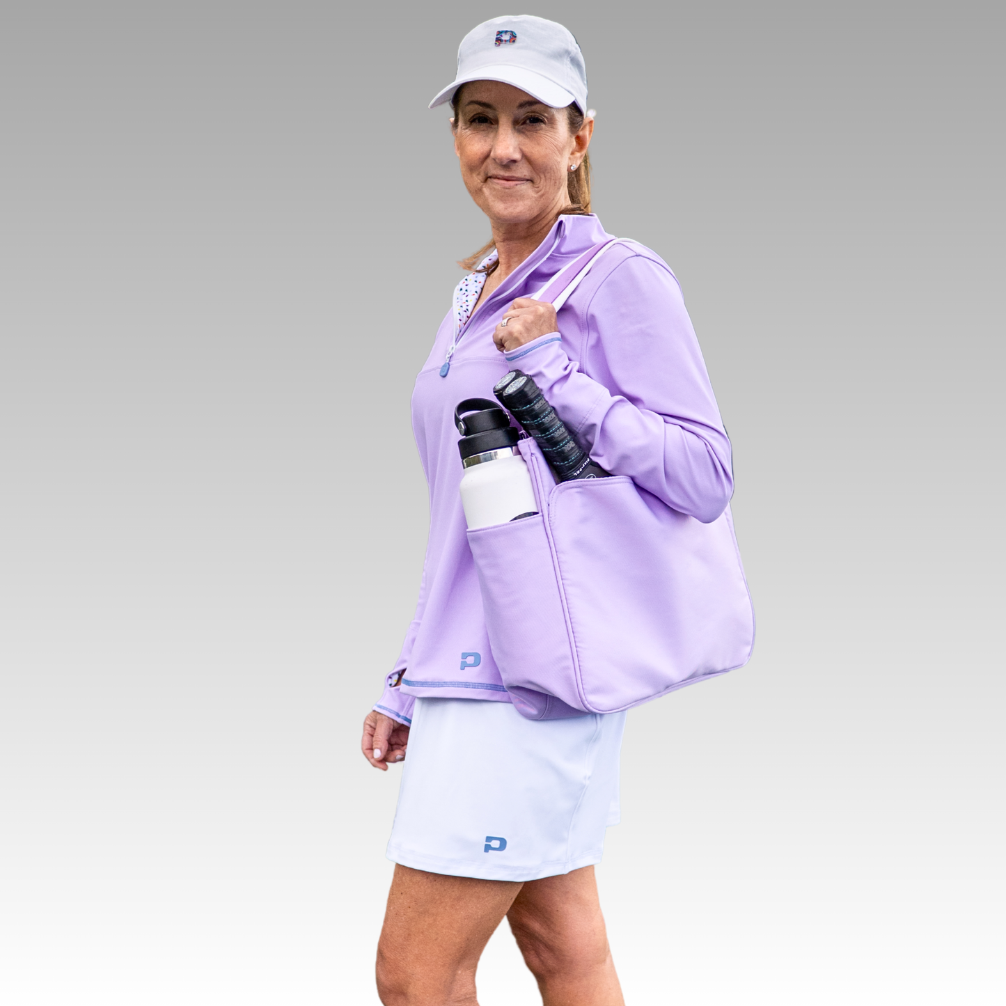 A woman in a lavender long-sleeve top, light blue skirt, and cap carries the Lulu Pickleball Tote by Playly Store, which holds a white water bottle and tennis racket. She stands against a gray background, smiling softly at the camera.
