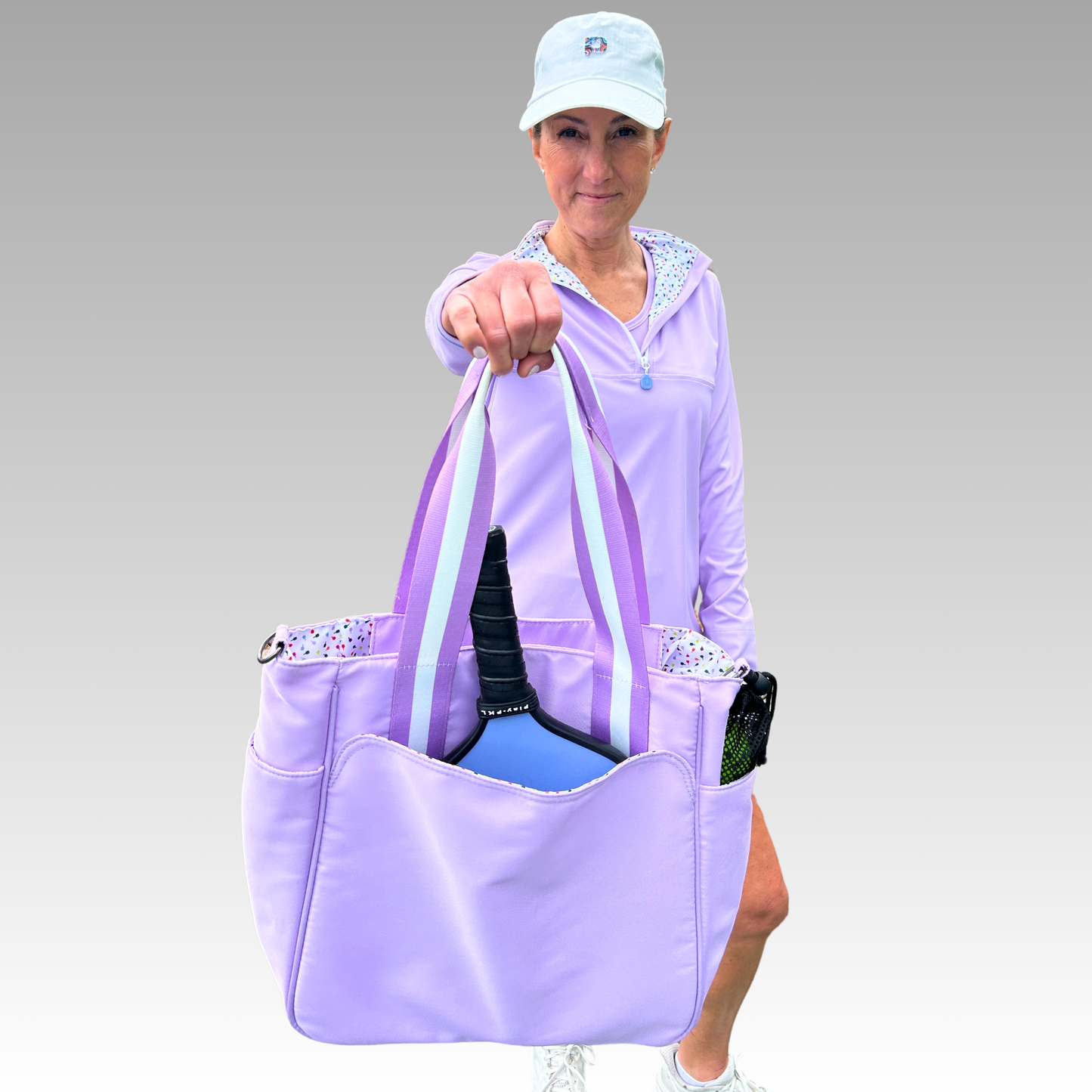 A woman in a light purple athletic outfit and blue cap confidently showcases the Playly Store Lulu Pickleball Tote, holding it toward the camera with a blue paddle and water bottle inside, set against a light gray gradient background.
