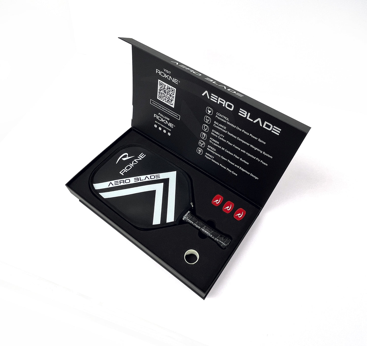 A black box with white text displays the ROKNE AERO BLADE 1.19 paddle, featuring a carbon fiber mono-frame and geometric design. The open box reveals the paddle, a metal accessory, and two red grip tapes neatly arranged in foam cutouts.