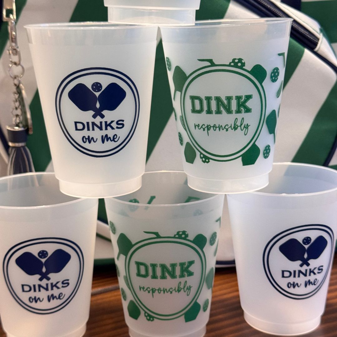 Five Fresh Pickle Pickleball Roadie Cups feature themed designs: three blue "DINKS on me" shatterproof cups and two green "DINK responsibly" cups, all stacked together with a green-and-white striped bag in the background.