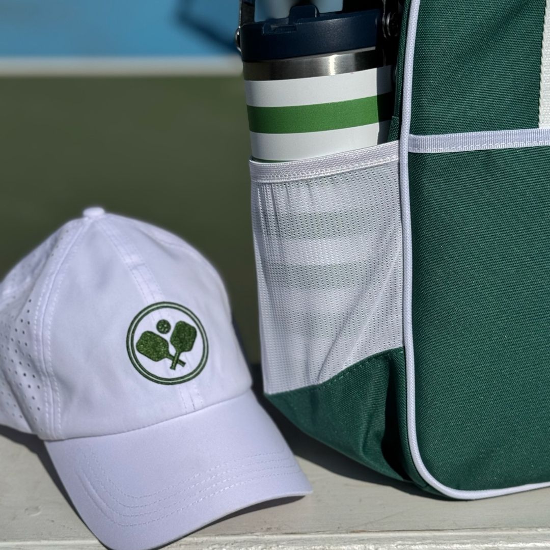 A white Fresh Pickle Pickleball Cap with a green logo rests next to a green-and-white tennis bag. A stainless steel bottle with green stripes peeks from the mesh pocket, set against a sunny, blurred tennis court backdrop.