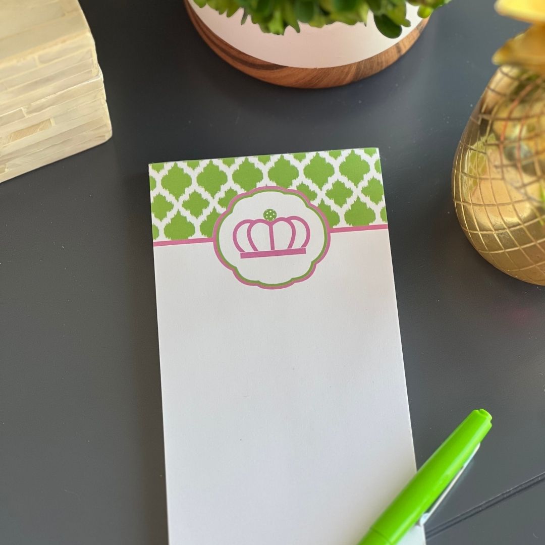 The Fresh Pickle Pickleball List Notepad, featuring a green and white top border and pink crown emblem, rests on a dark surface. A green pen, gold pineapple decor, and potted plant add flair—a must-have for any pickleball fan.