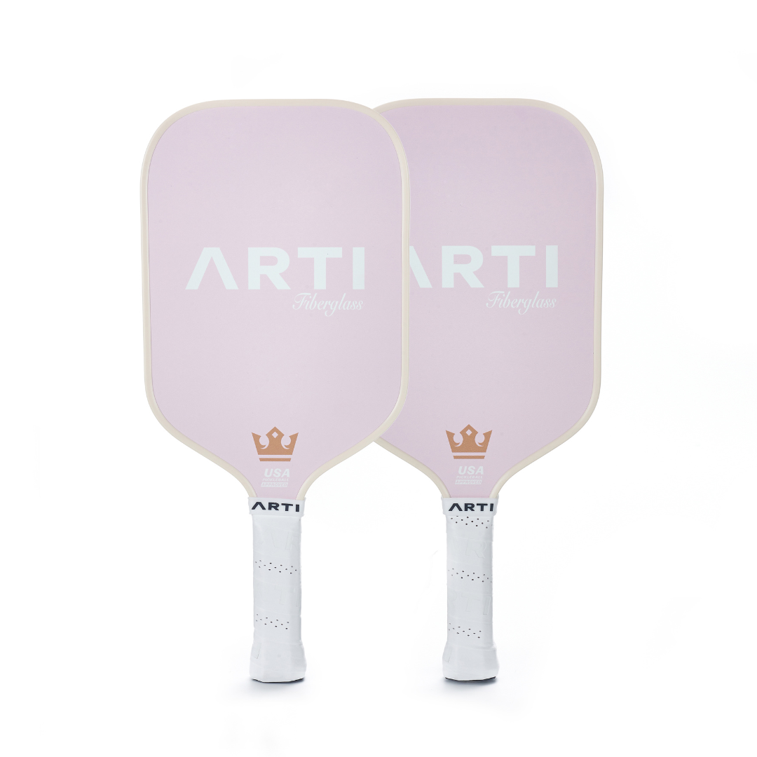 The ARTI Fiberglass "Valentine's" Pickleball Paddle Set features two pale pink paddles with white grips, bold white "ARTI" lettering, a gold crown and USA emblem, all from the brand ARTI, shown upright on a plain white background.