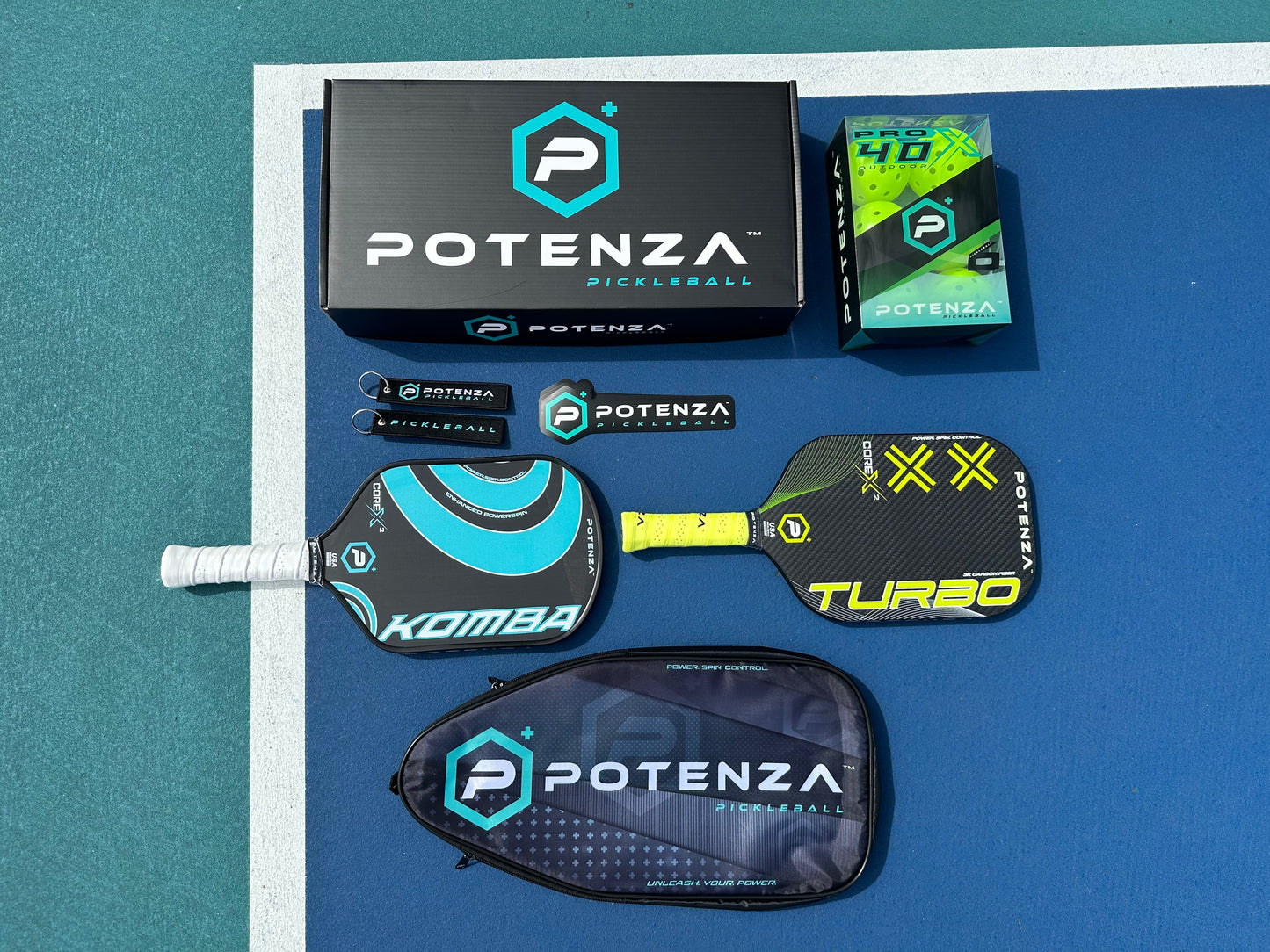 Two pro paddles, a POTENZA Paddle Travel Case (fits 2 paddles) by Potenza Pickleball, travel gear like a ball tube and keychains, Potenza pickleballs, and a large Potenza box are displayed on a blue-green court beside a white line.
