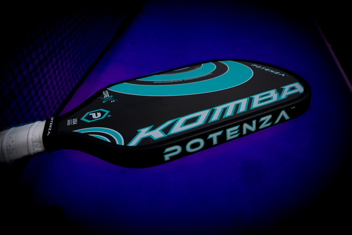 A close-up of the Potenza Pickleball KOMBA+ COREx2 paddle features a black, turquoise, and white design styled diagonally like premium carbon fiber. It stands out against a dark background with purple-blue gradient lighting and a blurred net.