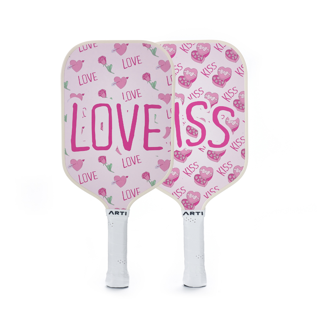 The ARTI Fiberglass "Valentine's" Pickleball Paddle Set features two pink paddles—one with “LOVE” and hearts, the other with “KISS,” lips, and hearts. Both have white handles, making this ARTI set perfect for couples.