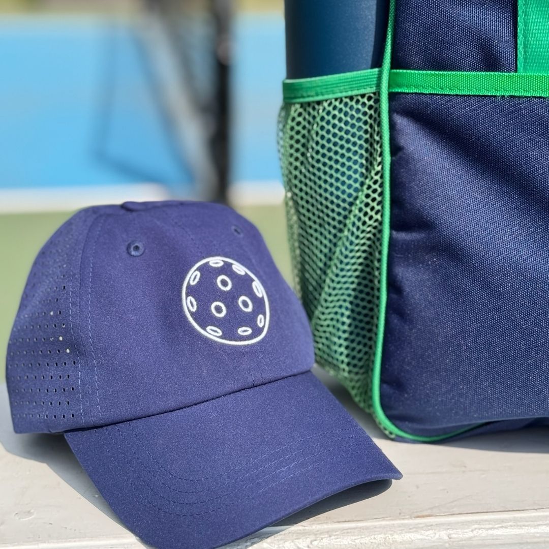 A Fresh Pickle Pickleball Cap in navy blue with a white pickleball graphic rests on a bench beside a navy and green sports bag with mesh pocket. The blurred background reveals a blue and green pickleball court.
