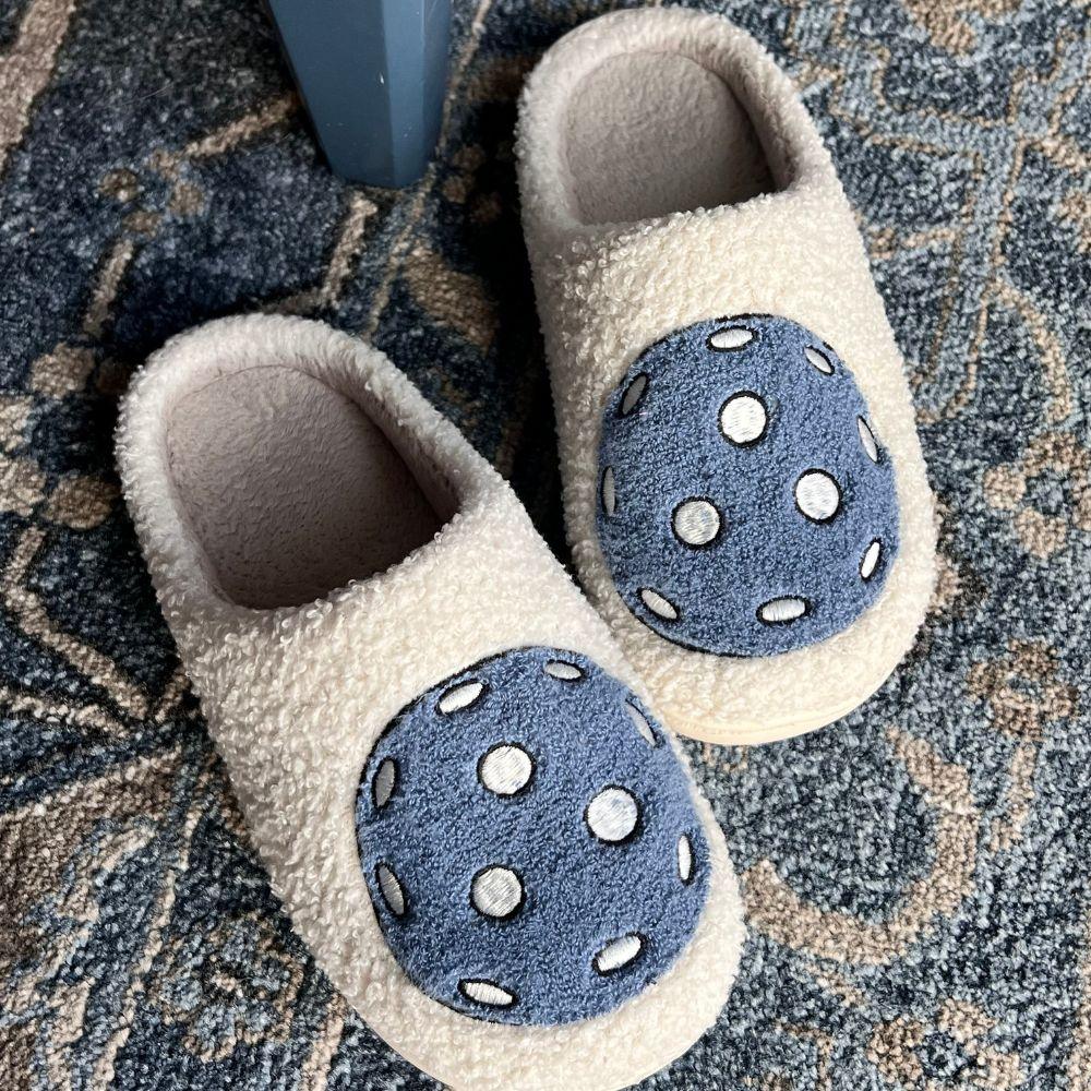 Slip into comfort with the Fresh Pickle Pickleball Slippers—off-white with textured blue circles and white dots—resting on a blue and beige carpet by a blue furniture leg. Perfect to pair with your pickleball pjs!.