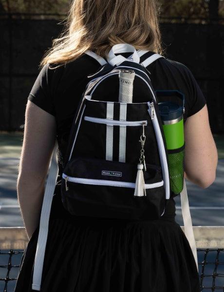 A woman stands by a tennis court fence with her back turned, wearing a black outfit. She carries the Fresh Pickle Teddi Pickleball Backpack, featuring white straps, zippers, and a roomy interior for her book and green water bottle.