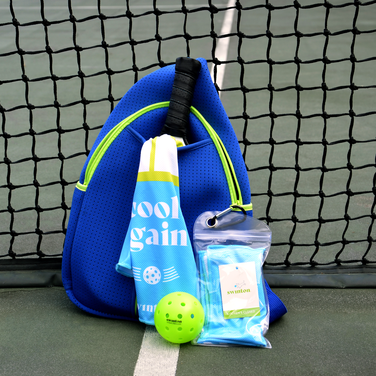 A blue zippered sports bag holds a tennis racket, Swinton Pickleball’s Pickleball Court Cooling Towel (blue, white, yellow), a neon green pickleball, and a clear pouch with gear—perfect for gifting on the pickleball court.