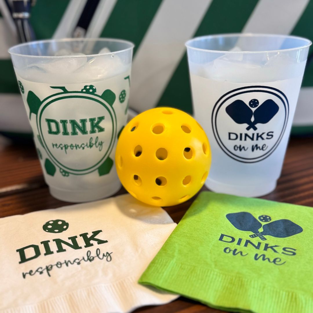 Two Fresh Pickle Pickleball Roadie Cups made from shatterproof plastic, filled with ice, sit next to a yellow perforated pickleball. One says “DINK responsibly,” the other “DINKS on me.” Nearby: matching napkins and a green-striped bag.