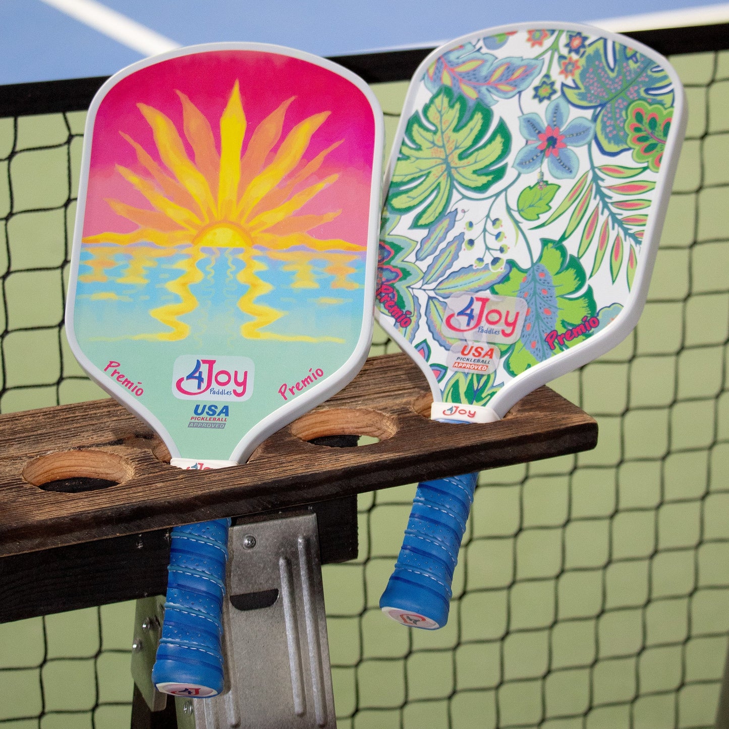 Two vibrant 4Joy Paddles Premio Series pickleball paddles with blue grips sit on a wooden holder by the net, showcasing high-performance composite construction; one features a sun over water, the other displays leafy florals.