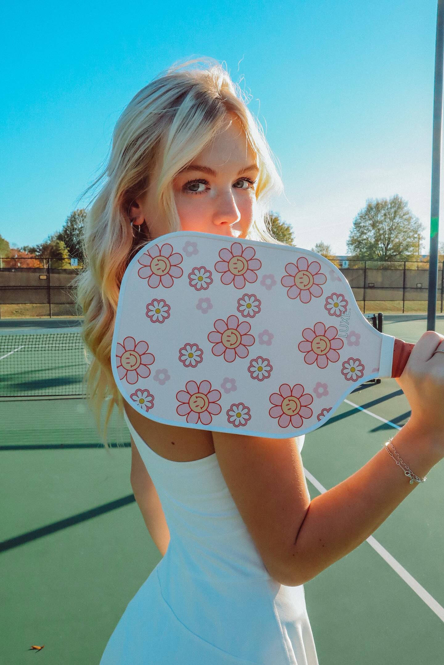 A woman with long blonde hair in a white dress holds the LUXE Pickleball Smiley Flower paddle, featuring pink and yellow flower designs, over her shoulder on an outdoor court beneath a clear blue sky with trees and fencing in the background.