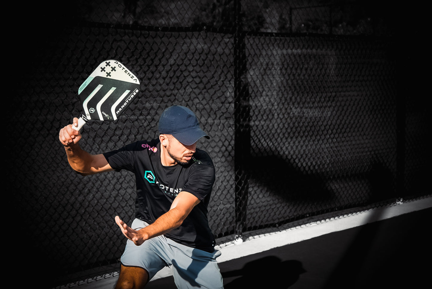 Wearing athletic gear and a navy cap, a man readies his swing with the Potenza Pickleball Carbon Nanotubes+ COREx3 paddle. Sunlight catches his focused look, while the court’s dark background adds dramatic emphasis to his powerful stance.