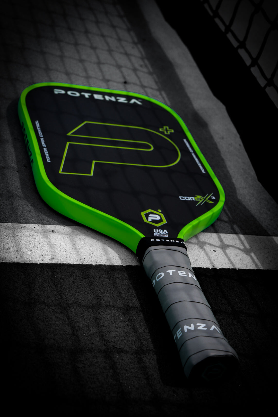 A Potenza Pickleball P+ PowerSpin Carbon COREx3 (Neon Green) paddle, featuring bold green edges, a large “P” logo, and a gray grip-wrapped handle, rests on an outdoor court near the net with its handle on the white line.