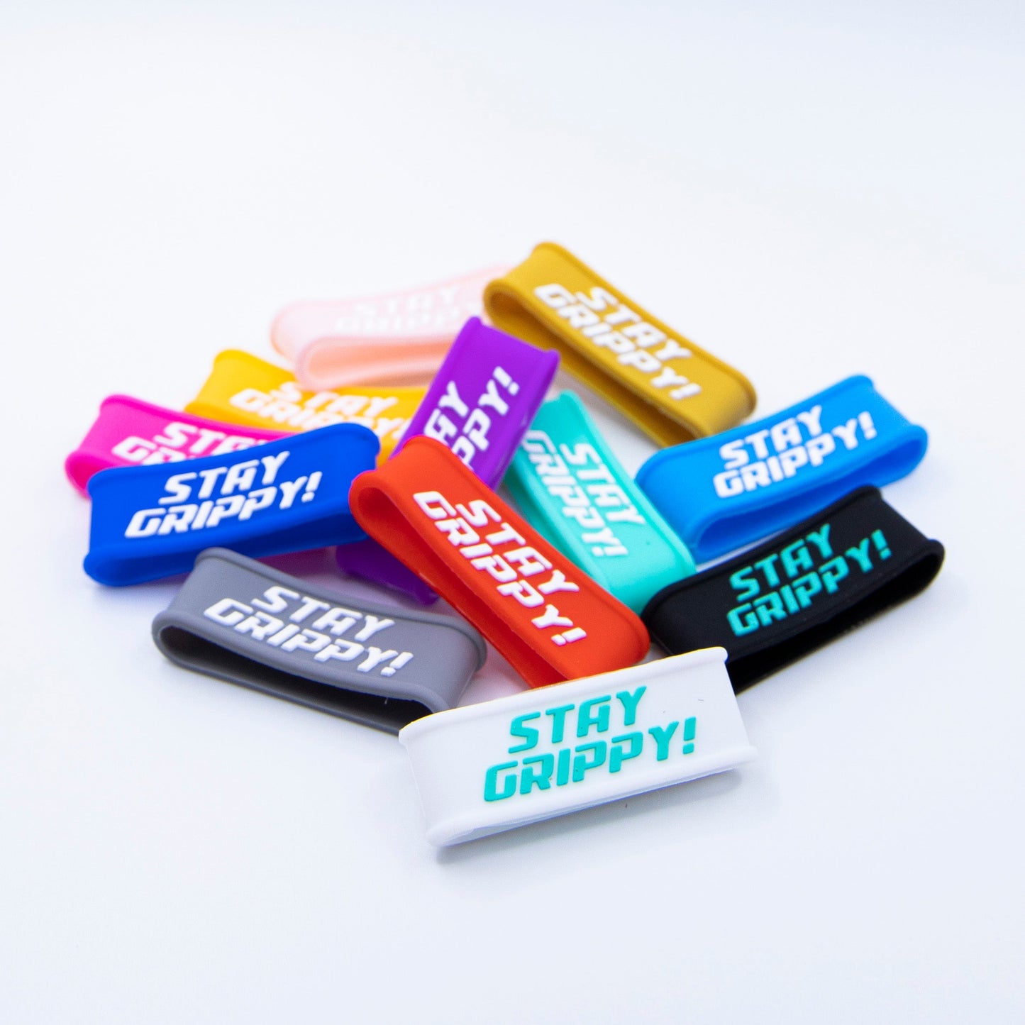 A colorful assortment of Bodhi Performance Paddle Bands lies scattered on a white background, each silicone grip accessory printed with “STAY GRIPPY!” in bold text and available in red, blue, pink, black, orange, yellow, gray, and white.