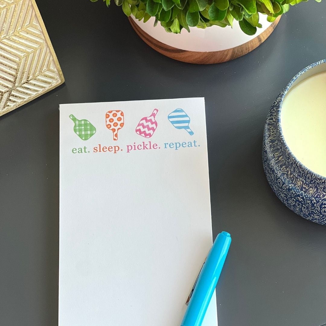 The Fresh Pickle Pickleball List Notepad, featuring colorful paddle icons and "eat. sleep. pickle. repeat." rests on a dark surface with a blue pen, potted plant, gold object, and blue-and-white candle nearby—perfect for any pickleball lover.