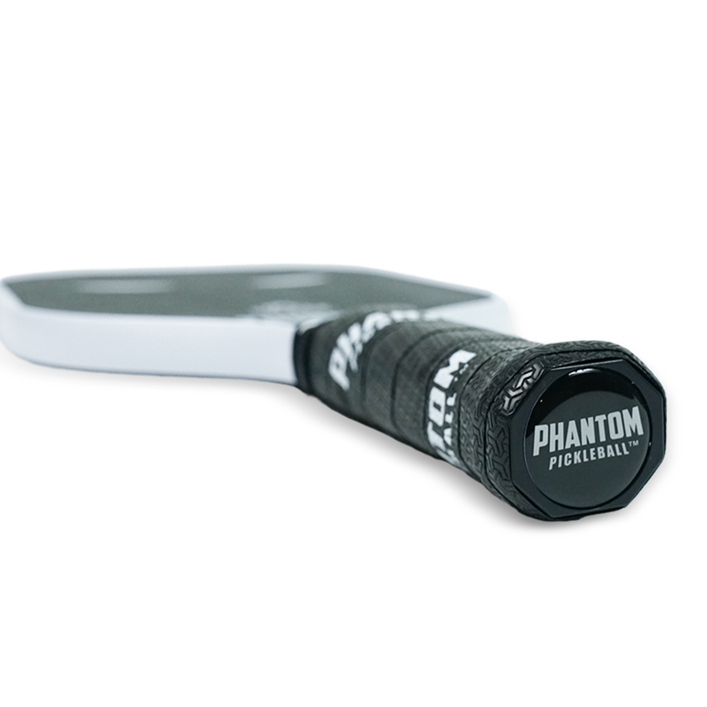Close-up of the Phantom Pickleball paddle handle reveals "Phantom Pickleball" on the end cap and grip. The Trinity 14MM T700 Raw Carbon Fiber Pro Paddle features a textured black grip, white edge, and its face blurred in the background.