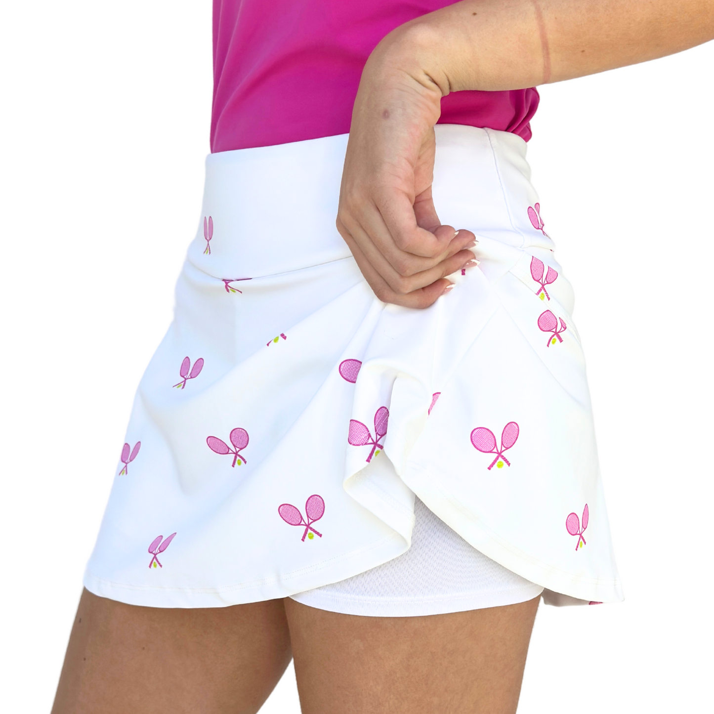 A model wears the Queen of the Court Love All Tennis Skirt with pink racket print, showing built-in white shorts, paired with a bright pink top. Made in USA. Only torso and upper legs are shown against a plain background.