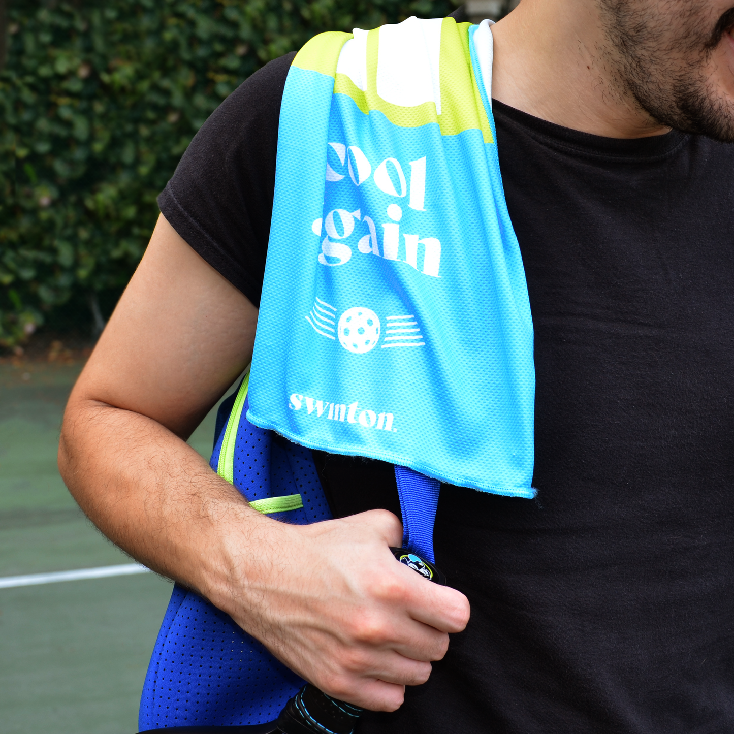 A person in a black shirt holds a blue sports bag and wears a Swinton Pickleball, Pickleball Court Cooling Towel (blue/yellow) reading "cool again" and "swinton" over their shoulder. Greenery and a pickleball court blur in the background.
