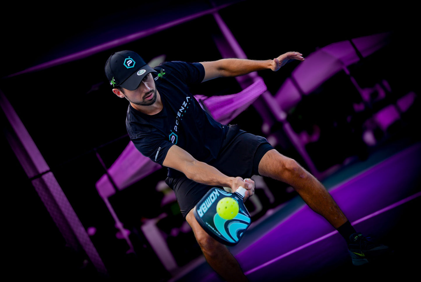 A man in athletic gear lunges on a pickleball court, intensely hitting a yellow ball with his Potenza Pickleball KOMBA+ COREx2 carbon fiber paddle. Vivid purple lighting accentuates his dynamic movement and the game's fast-paced action.