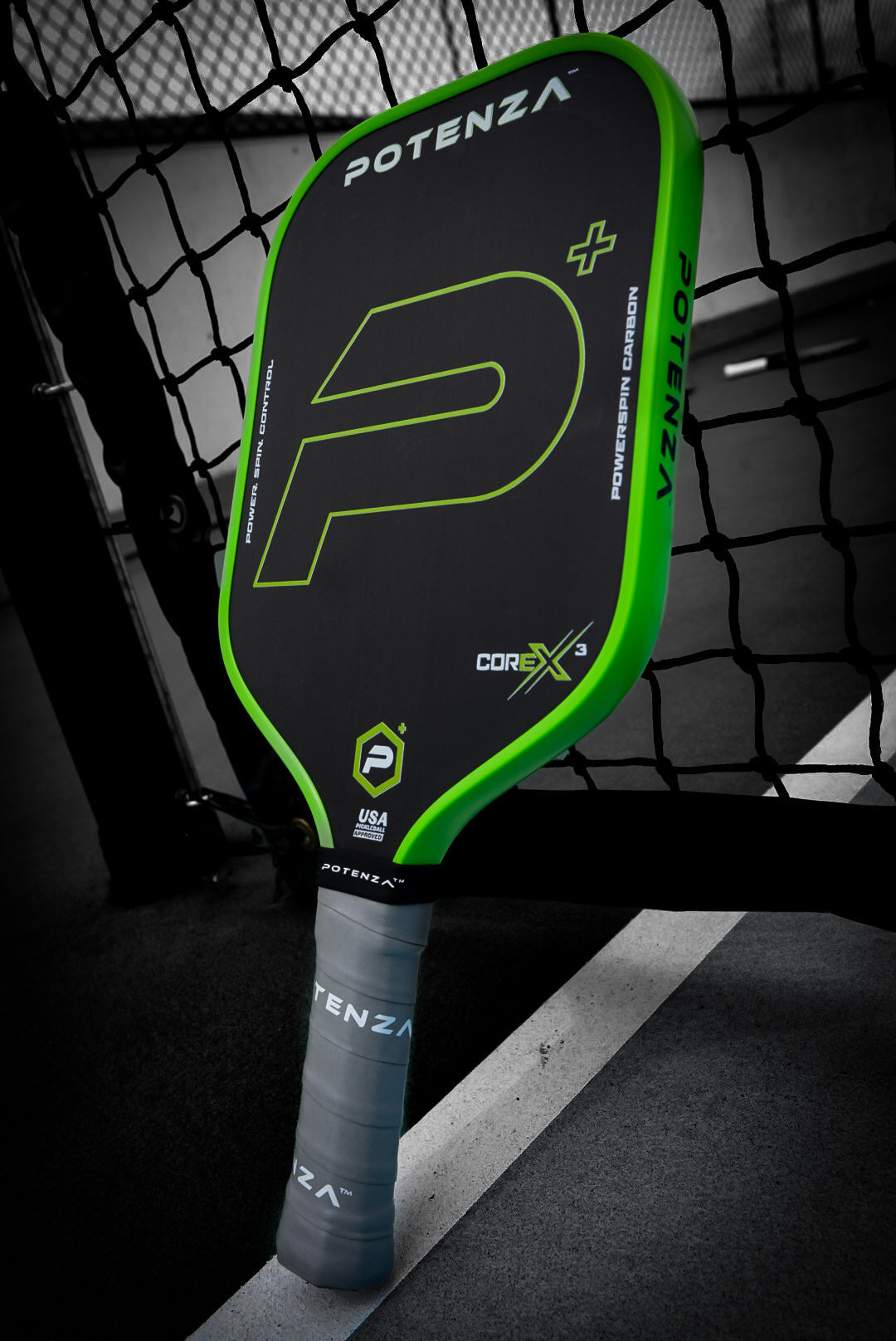 A Potenza Pickleball P+ PowerSpin Carbon COREx3 (Neon Green) paddle with black carbon fiber and green accents leans against a net on an outdoor court, showing the large P logo, CoreX branding, gray grip, and a chain-link fence in the background.