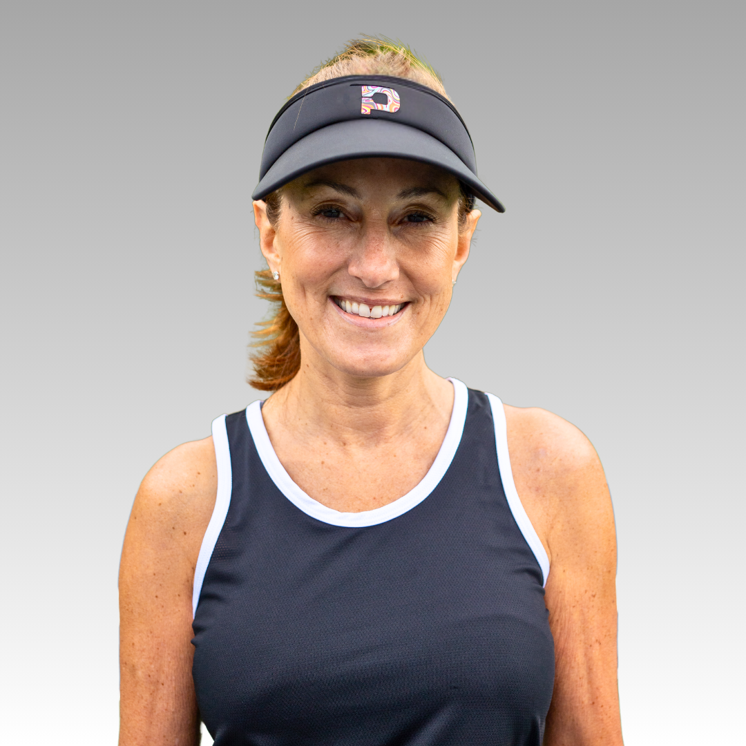 A middle-aged woman with light brown hair tied back smiles at the camera, wearing a black Playly Pickleball Visor by Playly Store and a navy blue sleeveless athletic top with white trim, set against a gray gradient background.