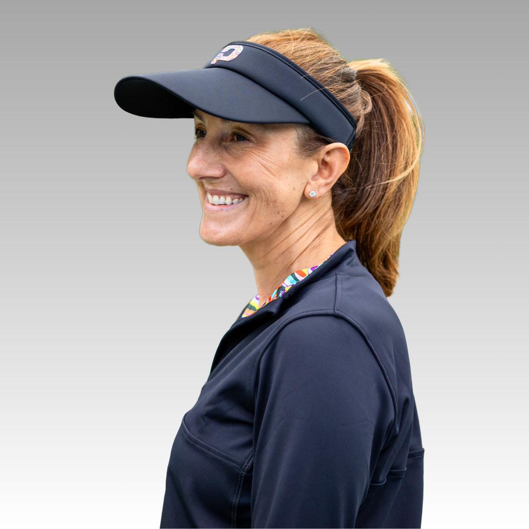 A smiling woman with light brown hair in a ponytail looks left, wearing a Playly Pickleball Visor by Playly Store, a black zip-up jacket, and a colorful collar. The background is a smooth gray gradient.