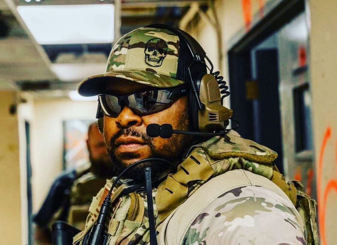 A man wears Epoch Eyewear Midway tactical sunglasses, a camouflage uniform, helmet with skull emblem, vest, and headset with mic while standing in an indoor corridor with blurred walls and red markings.