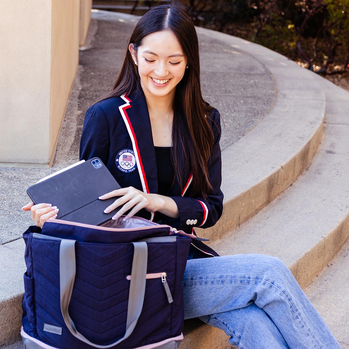 A young woman in a navy blazer and light blue jeans sits on concrete steps, smiling as she places a tablet into the chic Doubletake PARISII | Pickleball Tote on her lap. Bushes and a beige building appear in the background.