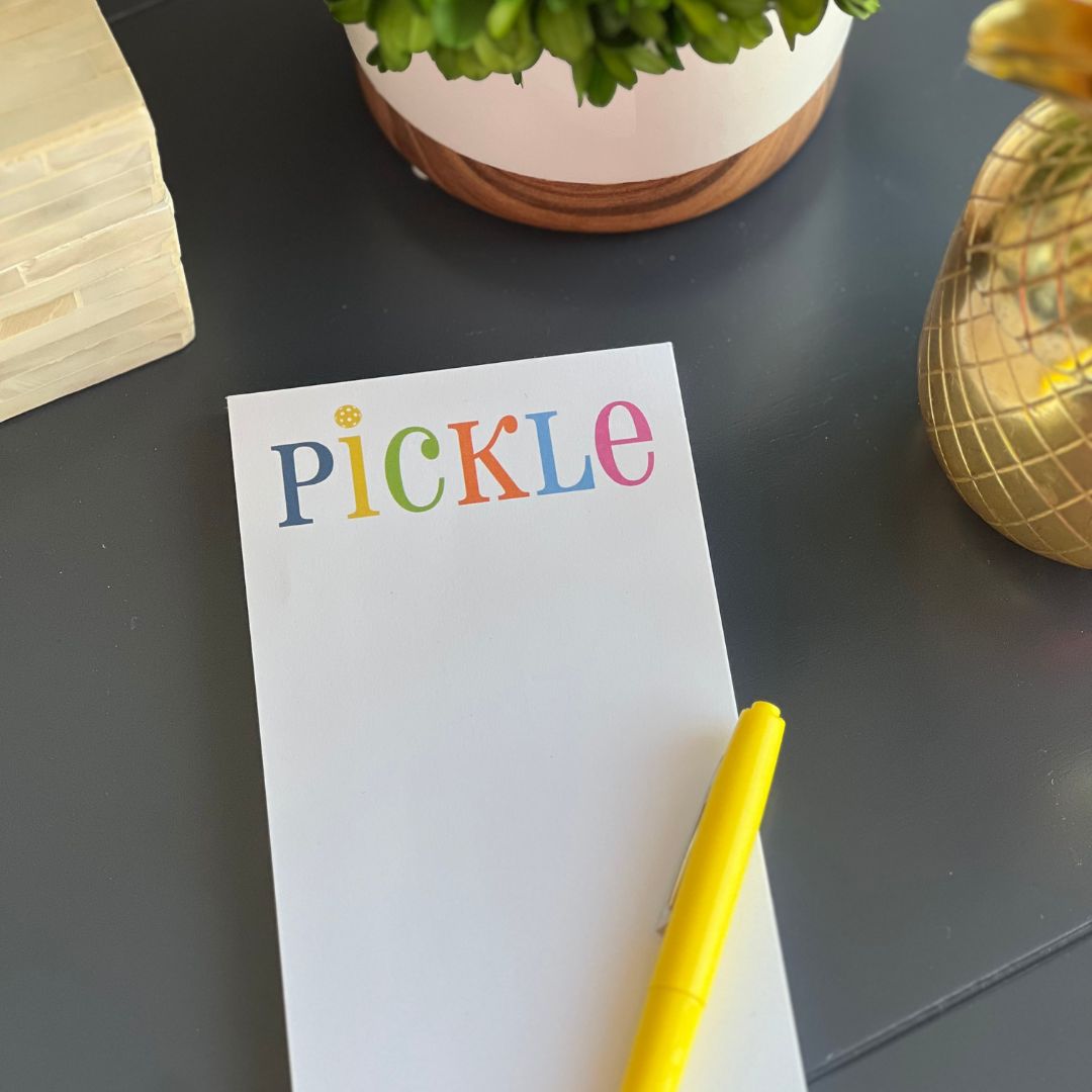The Pickleball List Notepad by Fresh Pickle features “PICKLE” in playful colors on the cover, paired with a yellow pen—perfect for pickleball fans. Styled on a dark table with a green plant, gold pineapple decor, and beige box.