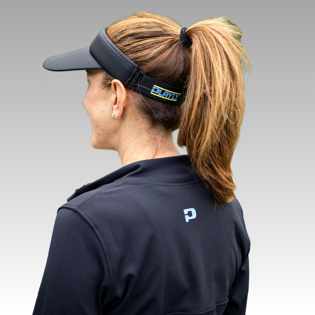 A woman with light brown hair in a ponytail wears the Playly Pickleball Visor from Playly Store and a black jacket with a blue P logo on the back. She is facing away, smiling, against a light grey gradient background.