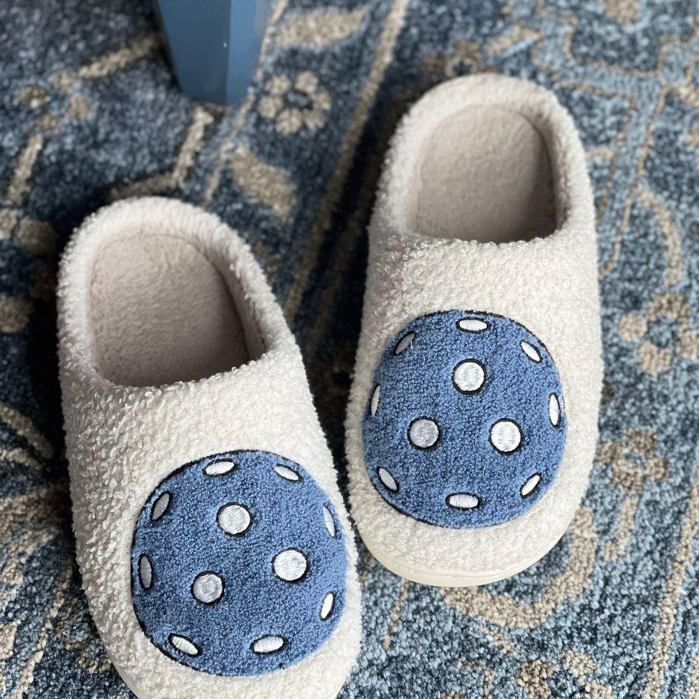 A pair of Fresh Pickle Pickleball Slippers in white with large blue polka dot circles are side by side on a blue patterned rug near blue furniture, featuring a cozy, textured design.