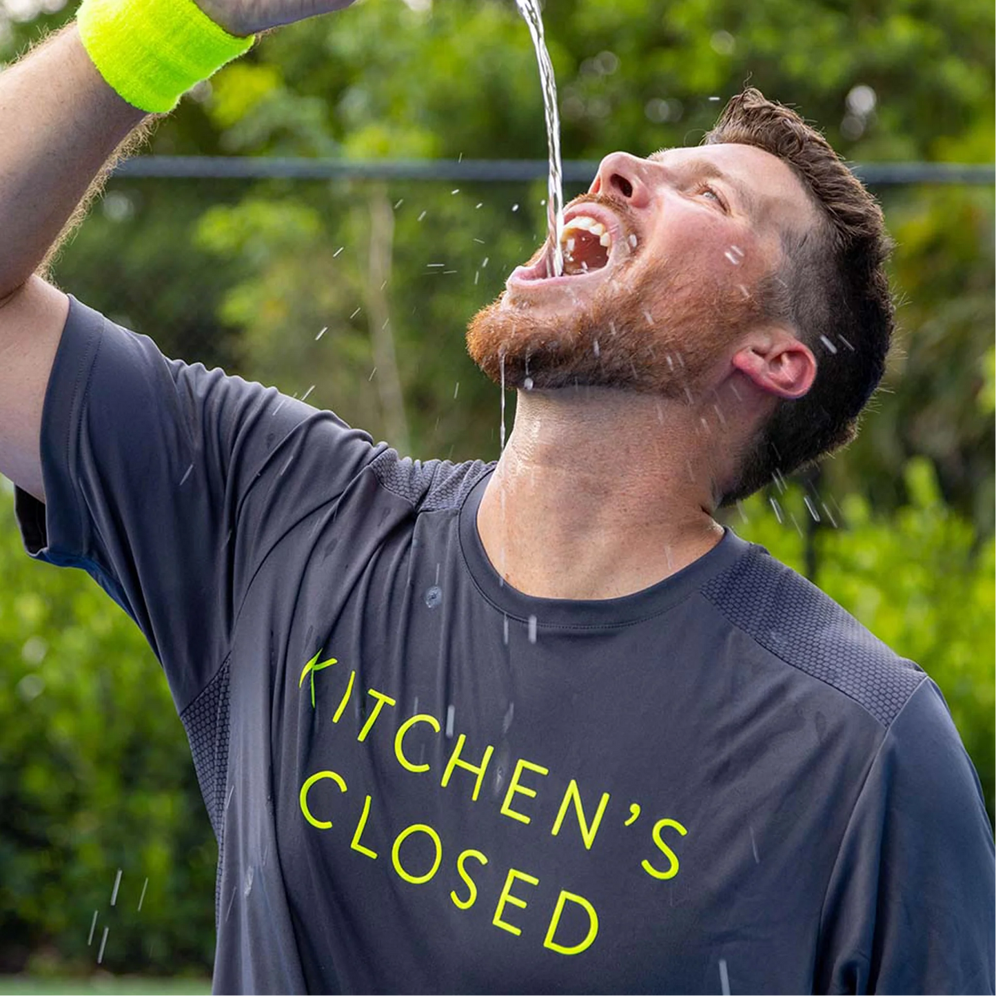 A man in a Swinton Pickleball Men's Kitchen's Closed Performance Shirt Gray, featuring bold yellow “KITCHEN’S CLOSED” text, pours water into his mouth by a pickleball court, sporting a yellow wristband.