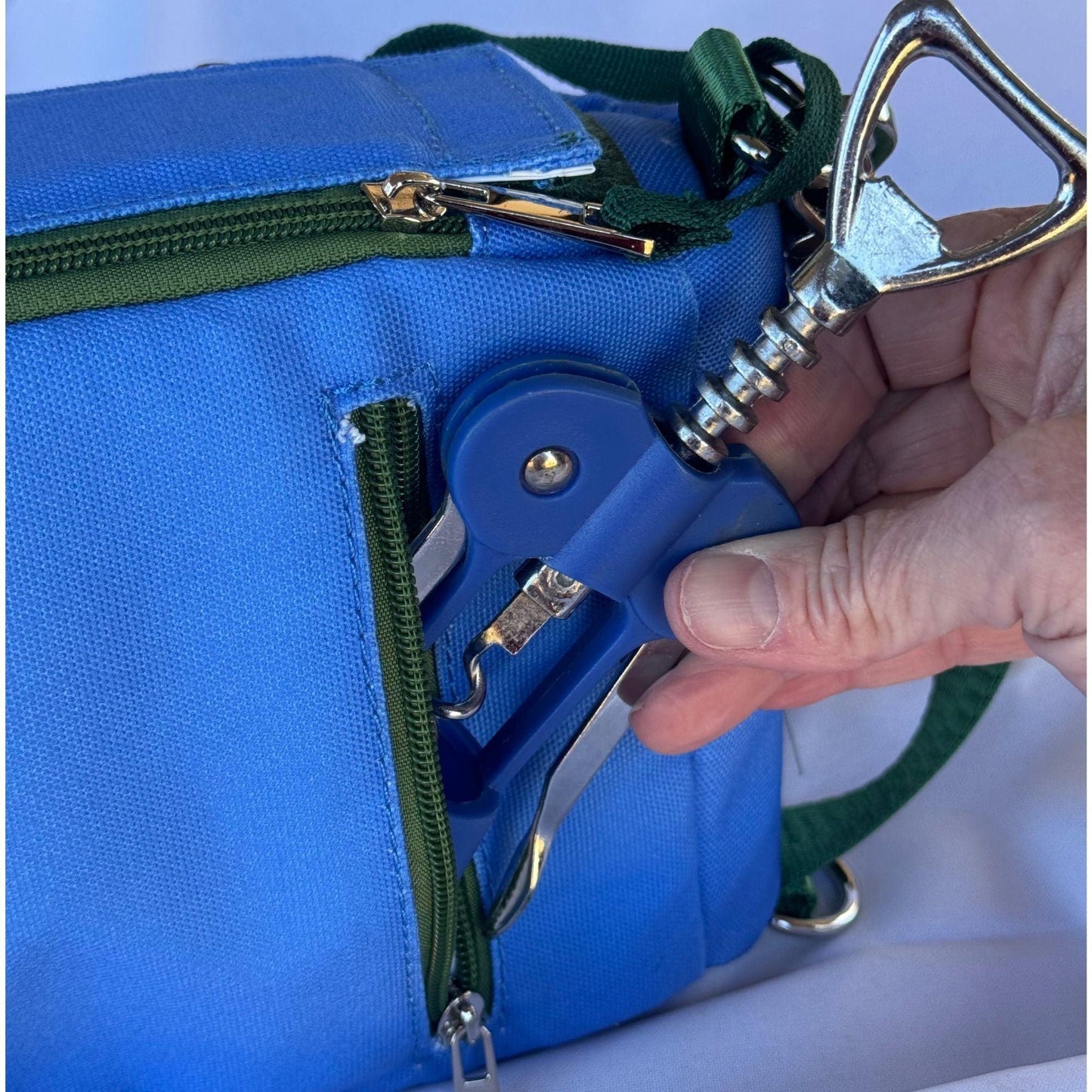 A hand pulls a blue and silver corkscrew from the green-zippered side pocket of a Fresh Pickle Insulated Cooler Bag with a shoulder strap, shown against a white background.