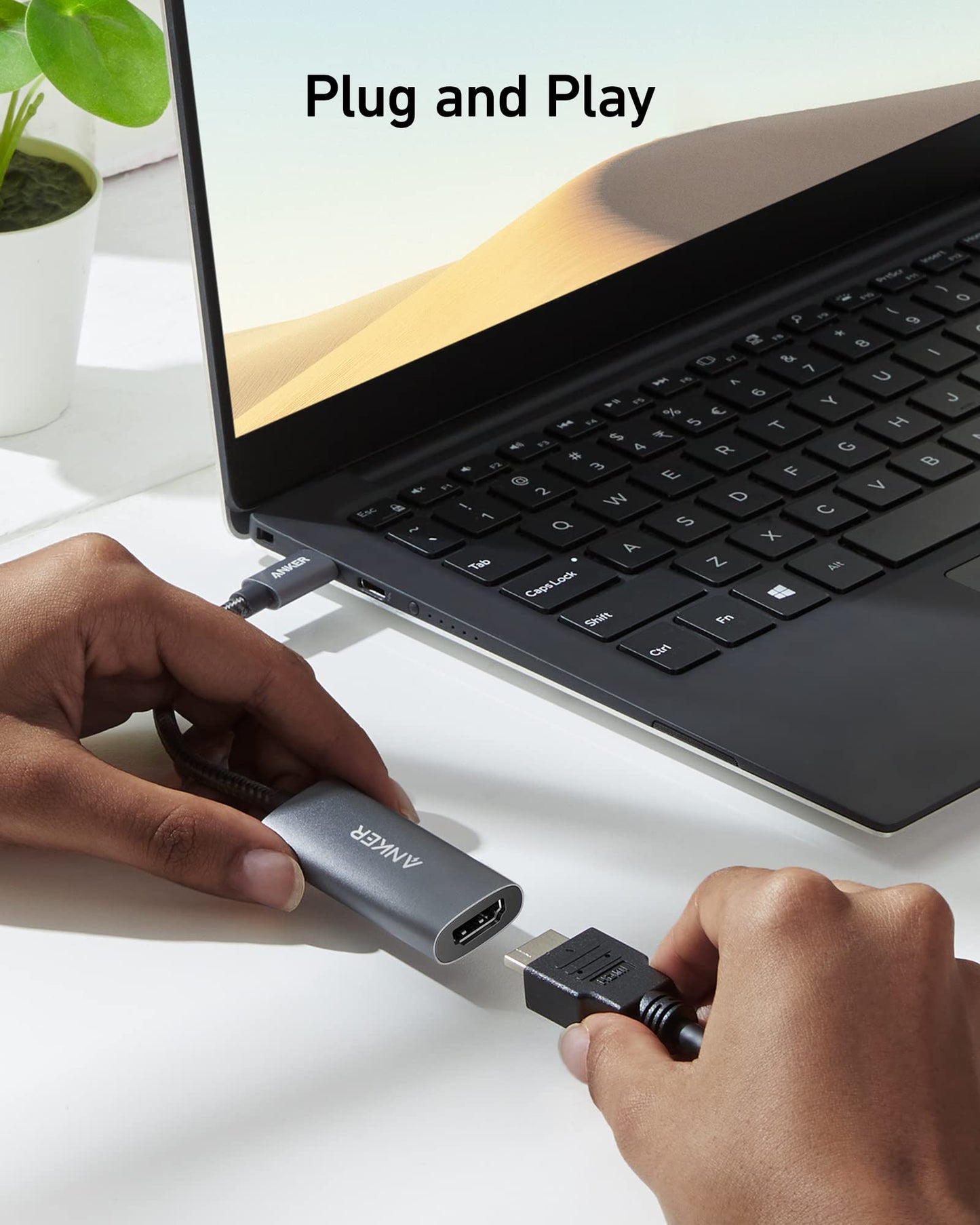 A person connects a USB-C to HDMI cable to the Anker 518 USB-C Adapter (8K HDMI) by Anker, attached to a black laptop on a white desk. A green potted plant is in the background. "Plug and Play" appears above the computer screen.
