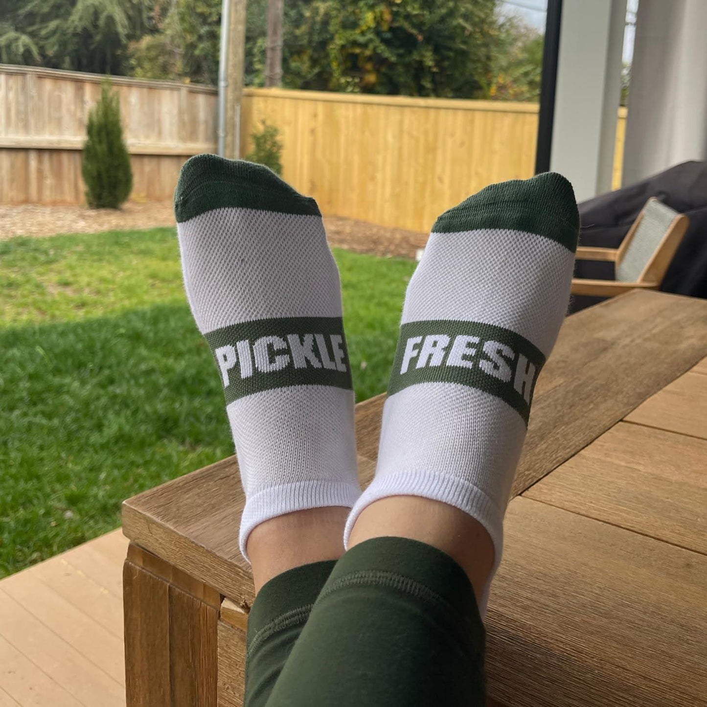 A person relaxes with feet up on a wooden table, wearing Fresh Pickle Pickleball Socks that say “PICKLE” and “FRESH,” with a grassy, fenced backyard in the background.