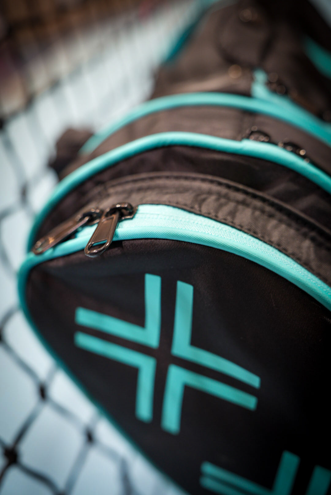 A close-up of the Potenza Pickleball Tournament Paddle Bag in black with teal zippers and geometric accents, resting on a surface before a blurred chain-link fence. The focus highlights its zipper, corner features, and premium paddle protection.