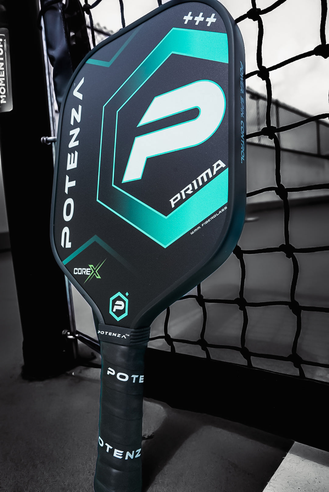 A close-up of the Potenza Pickleball PRIMA COREx paddle, featuring bold geometric graphics and “COREx” branding in black and teal, rests against a net with blurred court lines in the background.