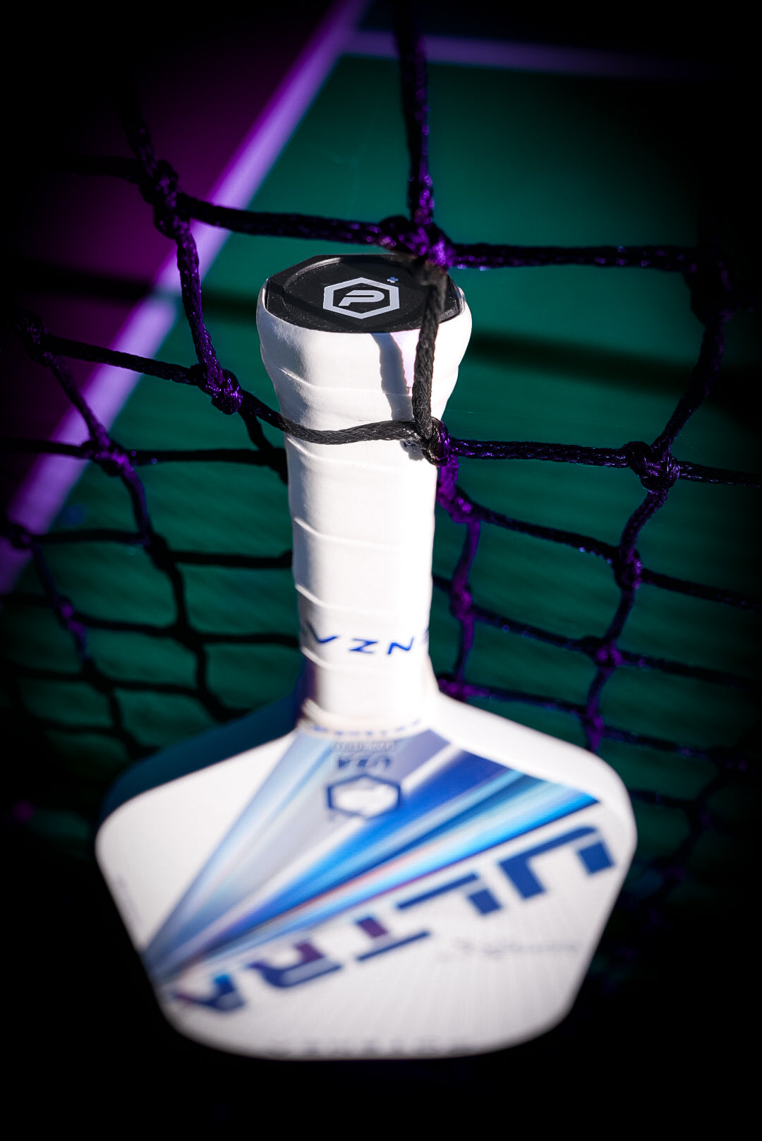 A white and blue Potenza Pickleball ULTRA+ COREx2 graphite paddle leans against a black net on a green and purple court. The white grip-wrapped handle is in focus, showing a hexagonal logo on the end cap.