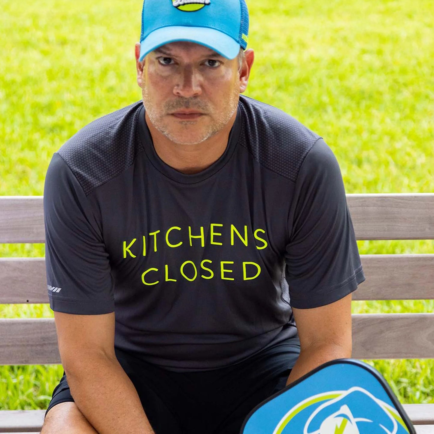 A man sits on a bench outdoors wearing a Swinton Pickleball Men's Kitchen's Closed Performance Shirt in gray. Sporting a blue cap, he holds a pickleball paddle and gazes seriously at the camera, with bright green grass behind him.
