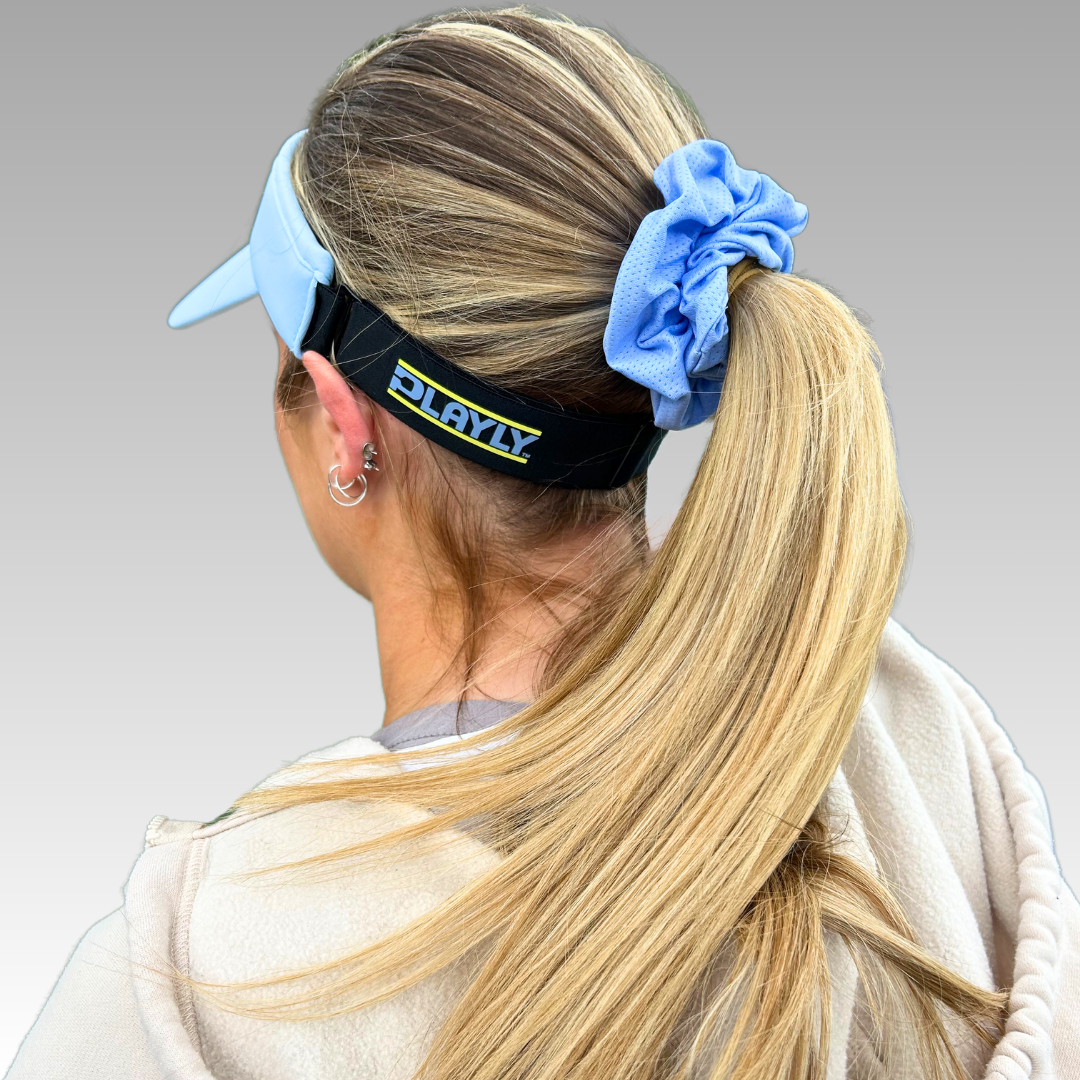 A woman with straight blonde hair in a ponytail wears a light blue Playly Pickleball Visor from Playly Store, featuring a black strap with yellow PLAYLY letters above her ears. She pairs it with a blue scrunchie and beige hoodie against a gray background.