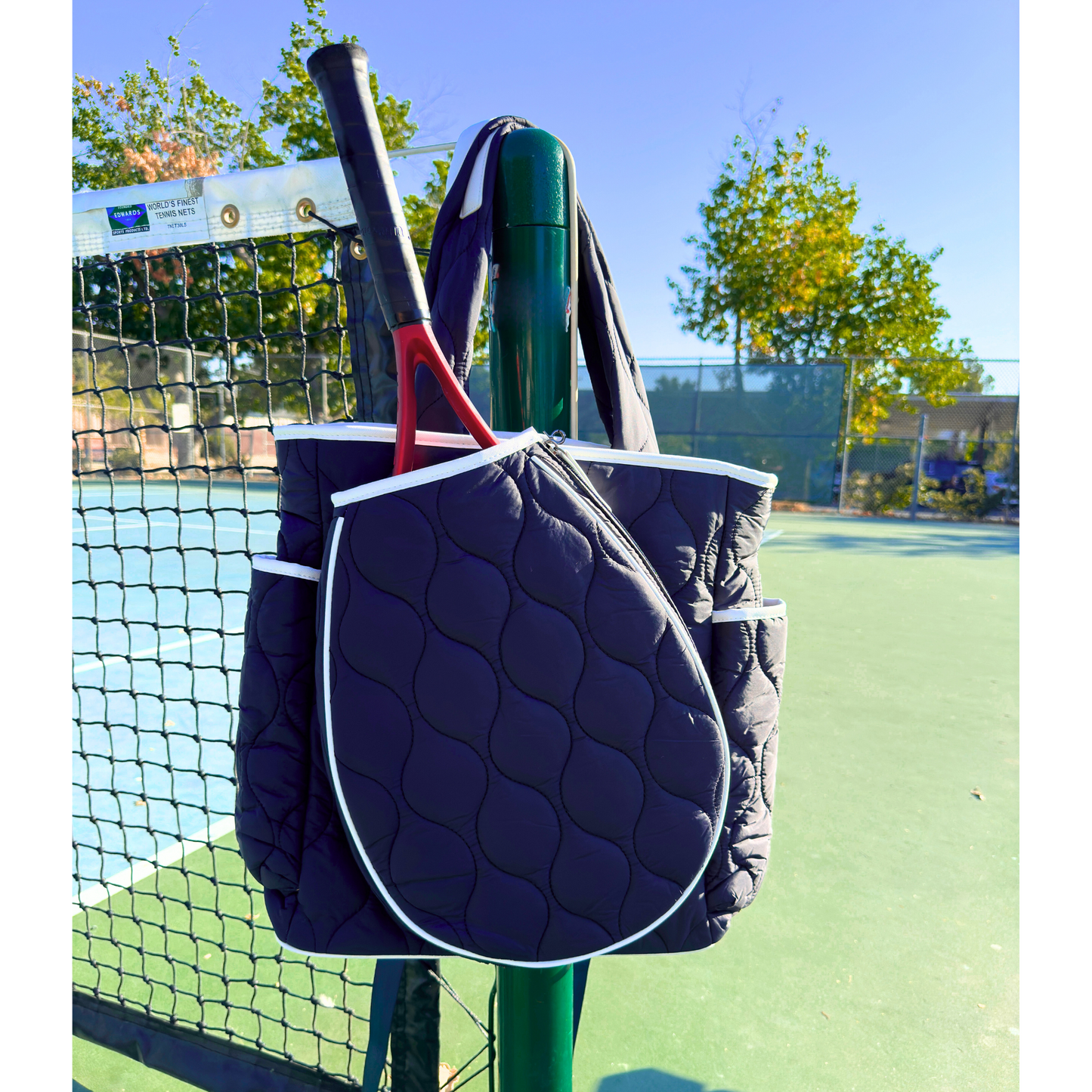 The Queen of the Court Park Ave Tennis Convertible Bag/Backpack in navy with white trim hangs on a green net post at an outdoor court, with a red and black tennis racket partially inside. The sunlit court, fence, and trees are visible in the background.