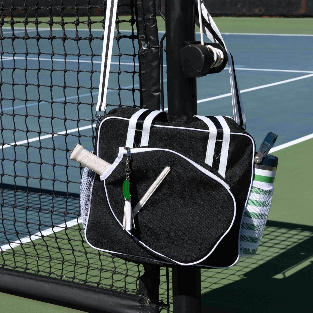 The Fresh Pickle Lola Pickleball Tote Bag in black and white hangs on a tennis net post. It features white trim, a racquet handle peeking out, a striped water bottle inside, and small accessories on the zipper pull.