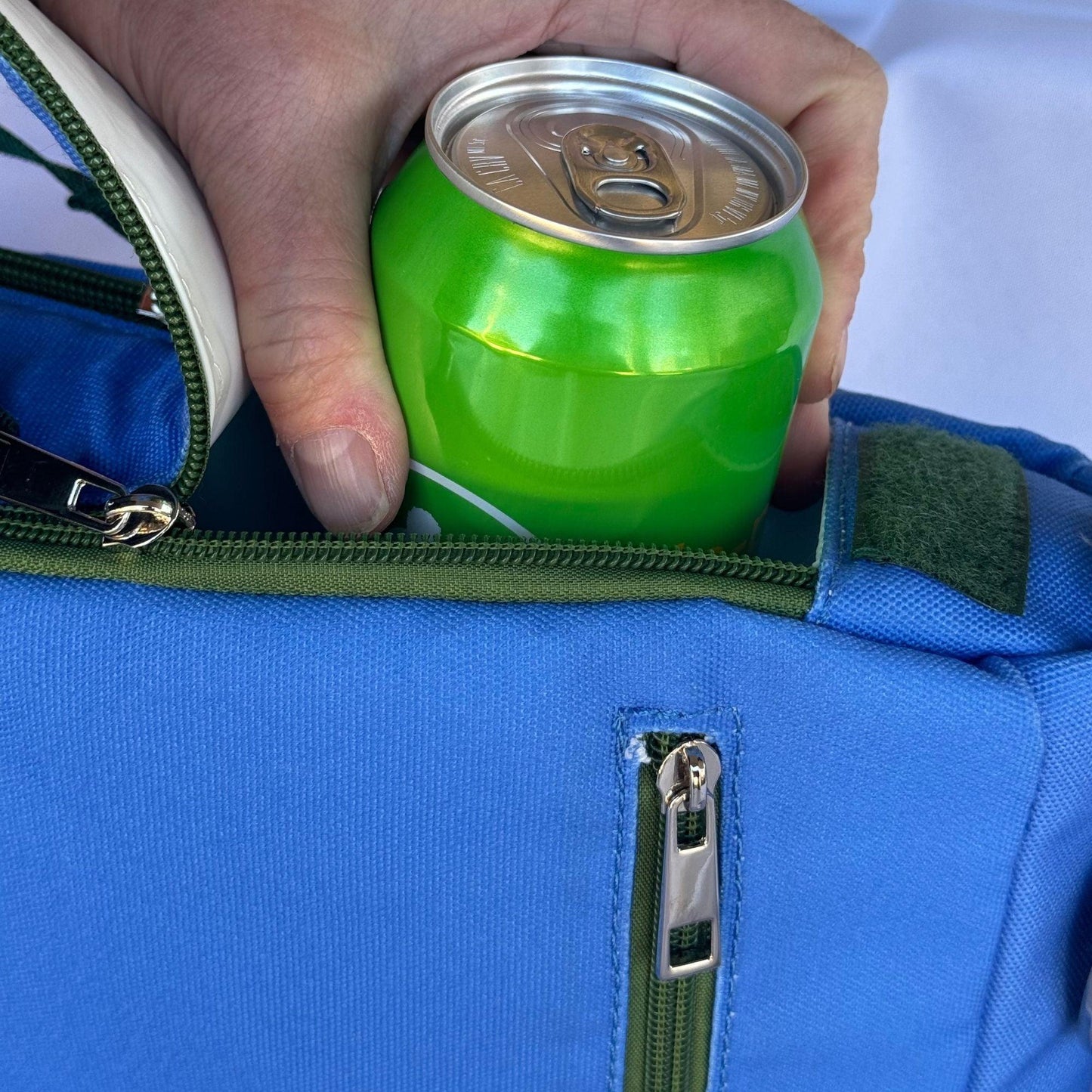 A hand places a green soda can into the Fresh Pickle Insulated Cooler Bag—a blue, soft-sided bag with a green zipper and a small front pocket—against a light background. The unopened can highlights its utility for carrying court necessities.