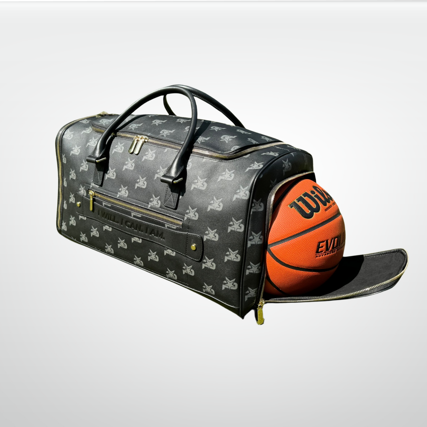 The iReliev I AM - Leather Monogram Training Duffel Bag in black rests on a light surface, decorated with white monogram logos. Its side pocket is open, showing a Wilson Evolution basketball. The bag features gold zippers and black handles.