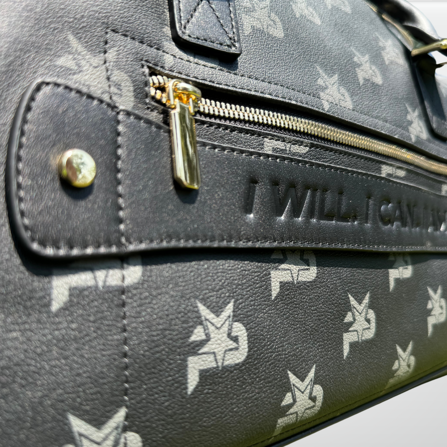 Close-up of the iReliev I AM - Leather Monogram Training Duffel Bag in black, featuring a repeating white logo, gold zipper, and an embossed “I WILL. I CAN.” leather strip that highlights the bag’s luxury texture and elegant details.