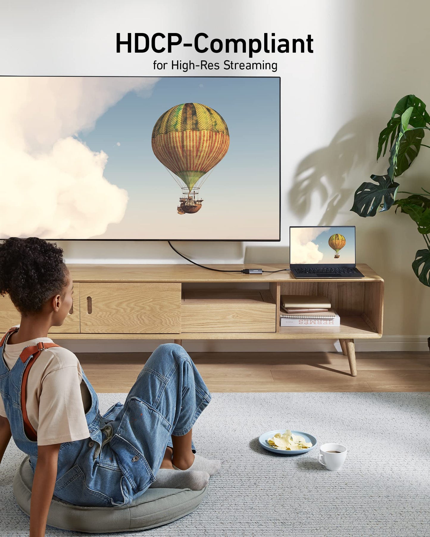 A person in a blue jumpsuit over a white shirt sits on a cushion, watching a hot air balloon scene on both TV and laptop connected with the Anker 518 USB-C Adapter (8K HDMI). A plate of food rests nearby.