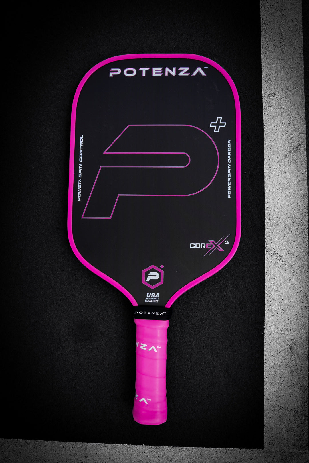 A black Potenza Pickleball P+ PowerSpin Carbon COREx3 paddle (Neon Pink, Elongated Face) with a neon pink edge and grip is centered on a dark background, featuring white and pink Potenza branding and a large stylized "P.