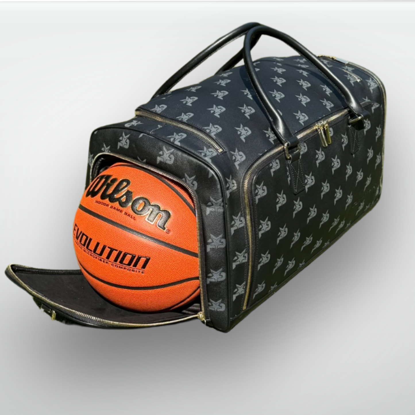The iReliev I AM - Leather Monogram Training Duffel Bag in black, featuring a repeating silver logo, is unzipped to show an orange Wilson Evolution basketball inside. The luxury bag sits on a light surface with its handles upright.